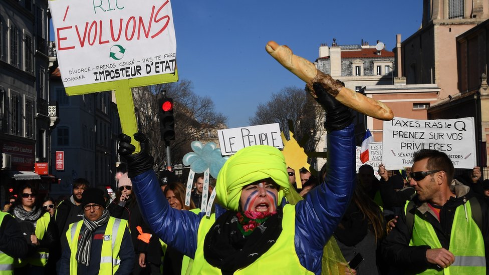 A protester wearing yellow vest (gilet jaune) holds a baguette and a sign reading "Citizens Referendum Initiative in Marseille, 15 December
