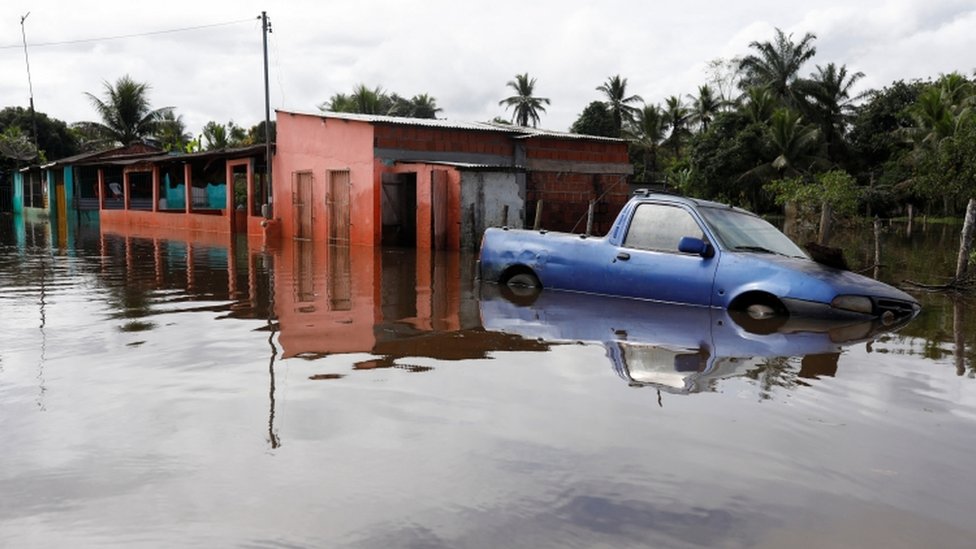 Carro em meio a água, com casas ao fundo