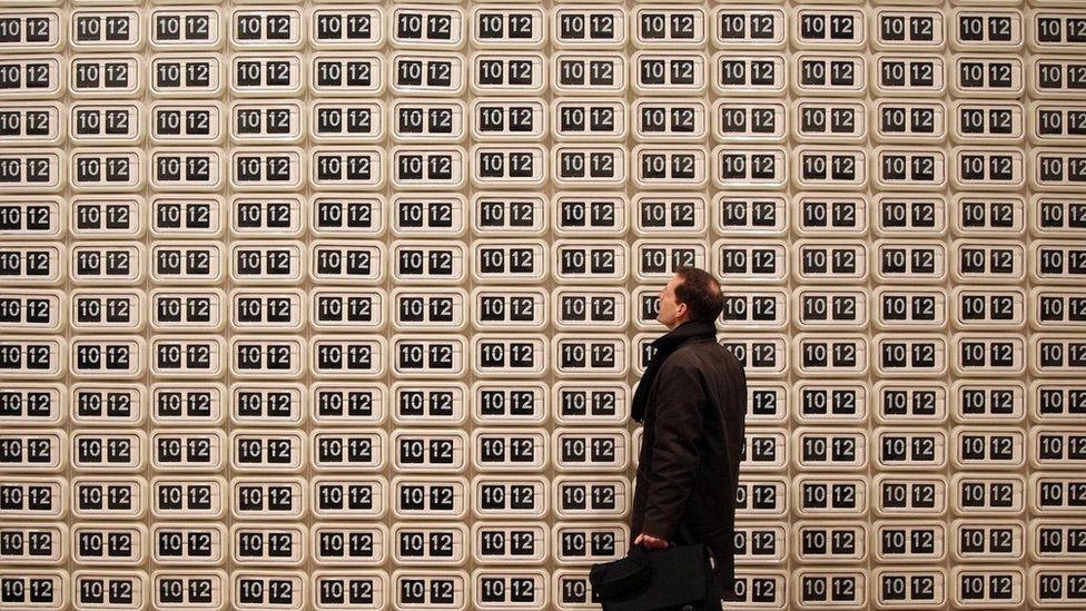 No somos muy buenos para darnos cuenta de cómo las decisiones sobre asuntos pequeños y cotidianos nos afectan en el largo plazo. Esto aplica al manejo de nuestro tiempo. Un hombre mirando una pared "empapelada" de relojes que marcan la misma hora