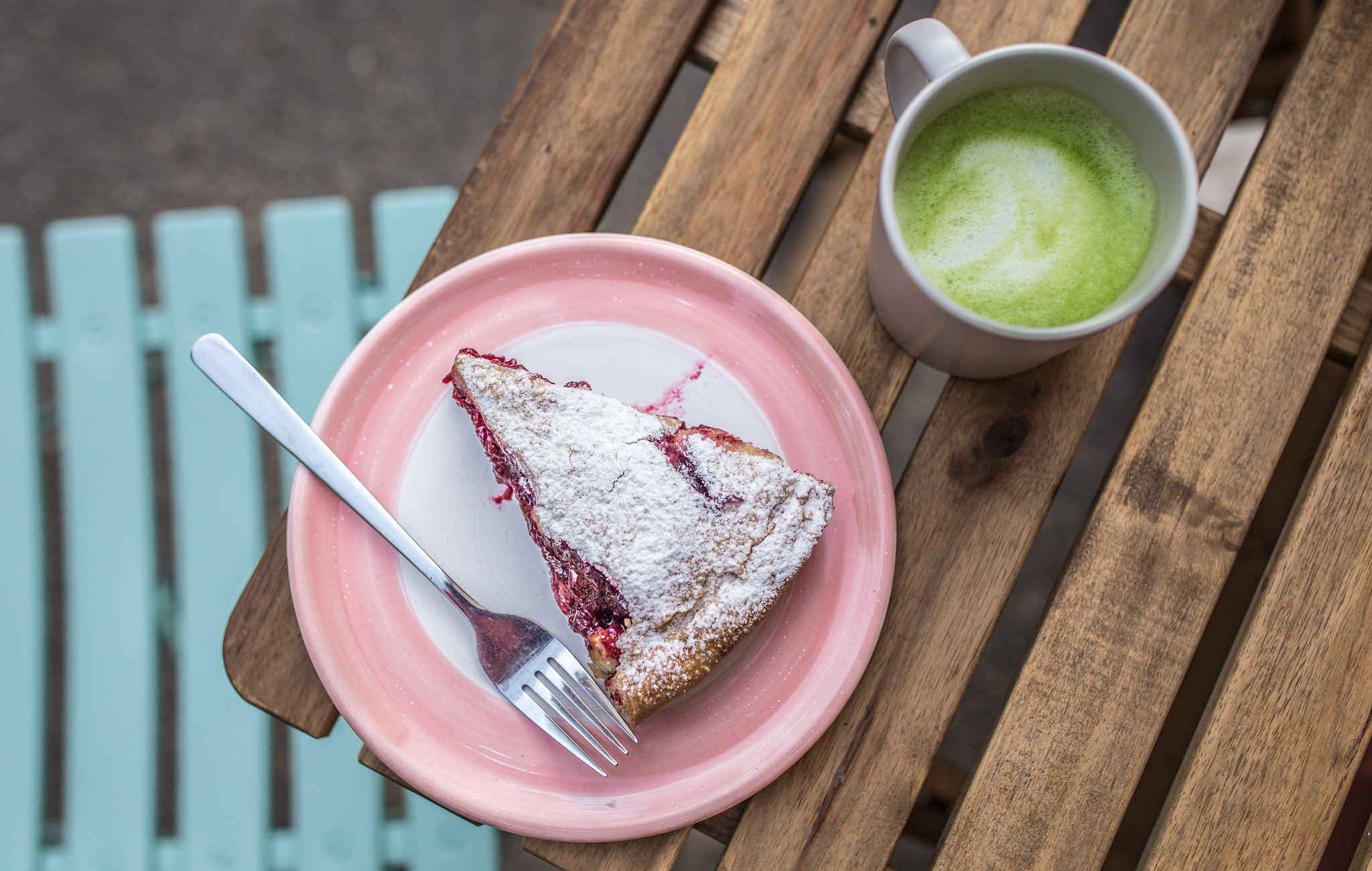 A cherry pie on a pink plate with a fork, and a cup of matcha latte, on a wooden table - photo is taken from above