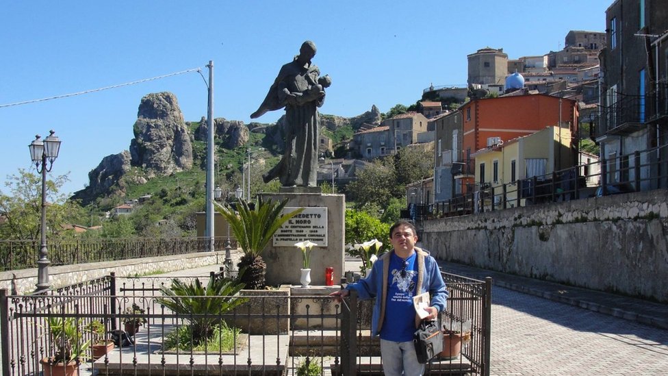 Pesquisador Lira, na Sicília, em frente a estátua em honra a São Benedito em praça sob dia ensolarado