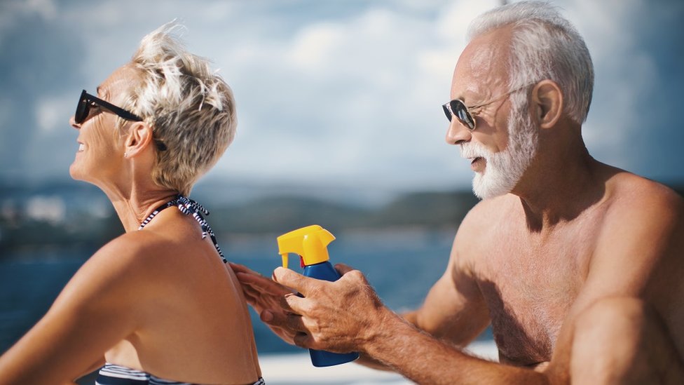 A senior couple sunbathing: the man is re-applying sunscreen on the woman.
