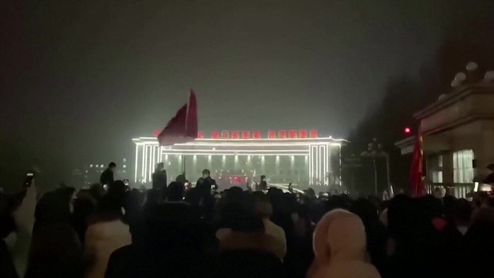 A large crowd of protesters is seen in front of an illuminated building in the Chinese city of Urumqi at night
