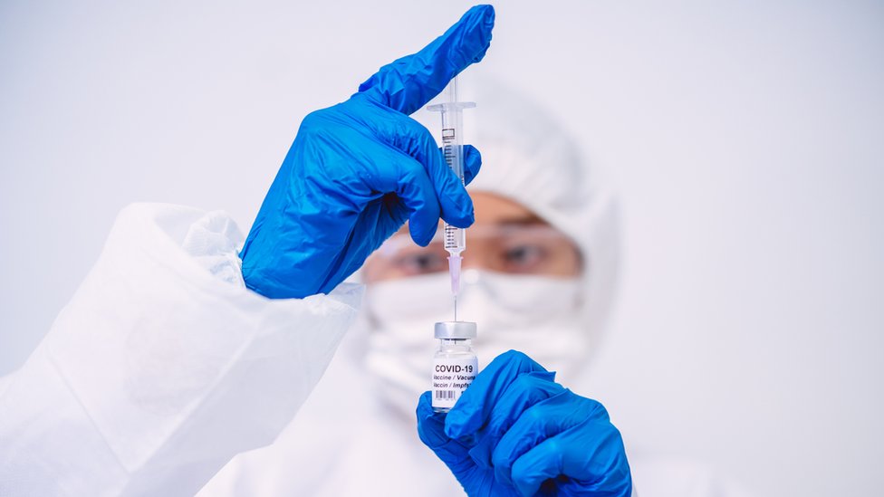 Врач в защитных перчатках и спецодежде заполняет шприц для инъекций вакциной COVID-19