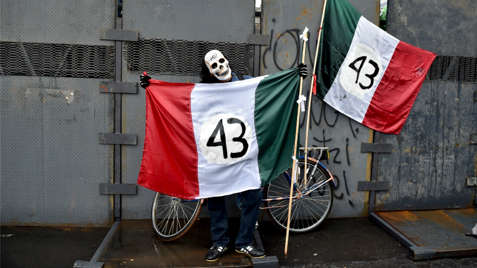 Una persona enmascarada sosteniendo dos banderas de México en las que se lee "43".
