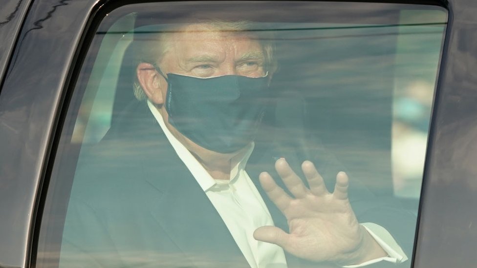 El presidente de Estados Unidos, Donald Trump, con una mascarilla y saludando con la mano desde un auto con la ventana cerrada