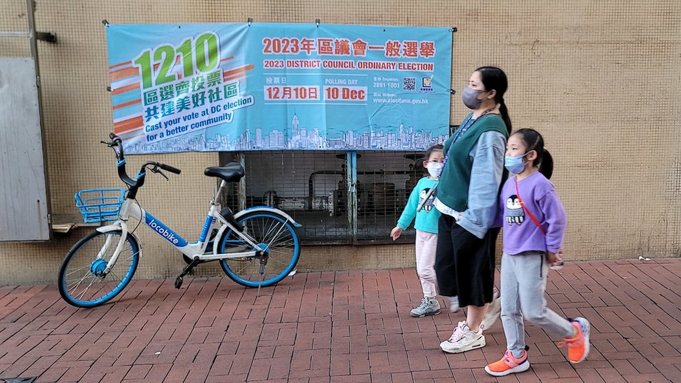 香港元朗街頭一位女士帶著小孩走過一幅政府宣傳區議會選舉的橫幅（BBC中文圖片19/11/2023）