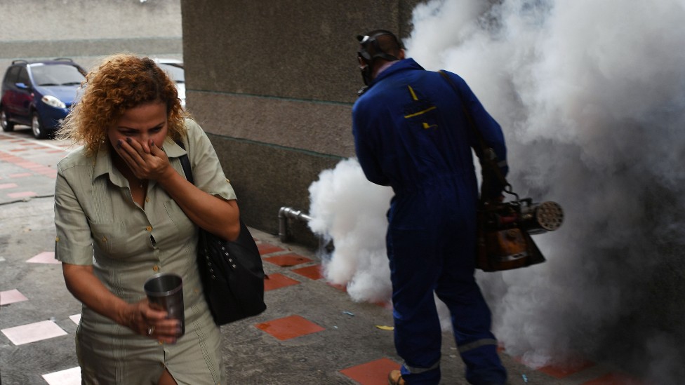 Un mujer se cubre la boca a pasar junto a un trabajador municipal que está rociando productos químicos para contener la propagación de mosquitos en Venezuela