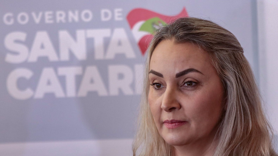 Daniela aparece de perfil, com painel atrás dizendo 'Governo de Santa Catarina'
