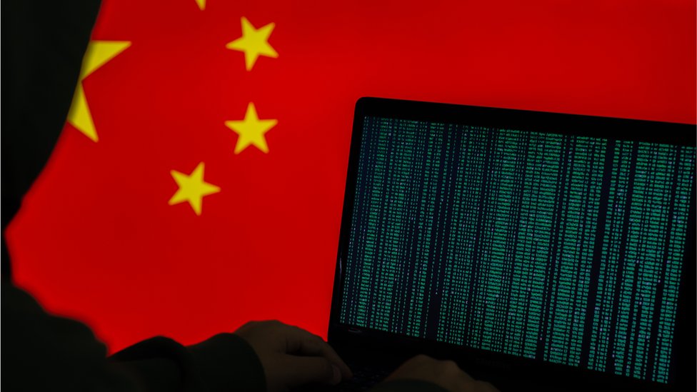Imagen de stock de una persona encapuchada que trabaja en la computadora frente a una bandera china.