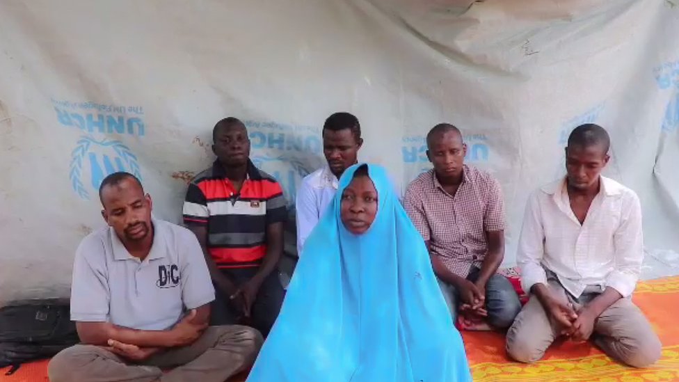 Кадр из видео, на котором запечатлены похищенные работники гуманитарных организаций и пятеро мужчин, предположительно ее коллеги. Все сидят на циновках перед брезентом.