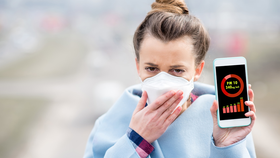 Mujere con una máscara y un celular que muestra el nivel de partículas gruesas de contaminación o PM10.