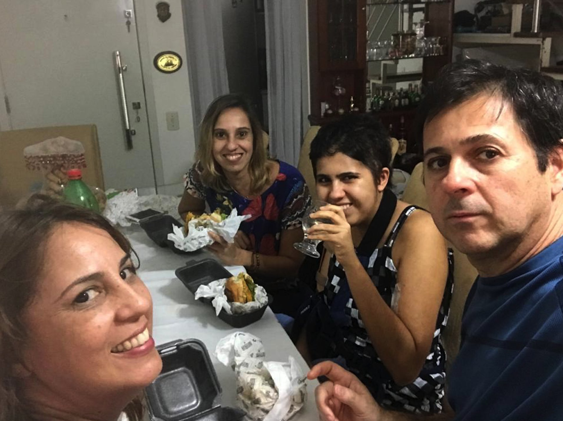 Selfie feita por Leila, ao lado de Uarlem. Ao fundo Larissa e a tia. Eles estão comendo um sanduíche.