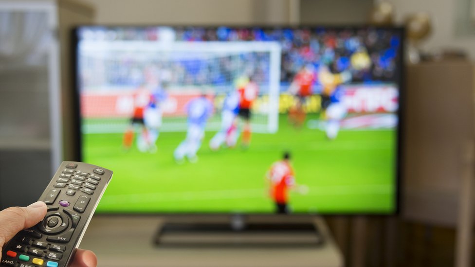 Controle remoto com televisão passando futebol desfocada ao fundo