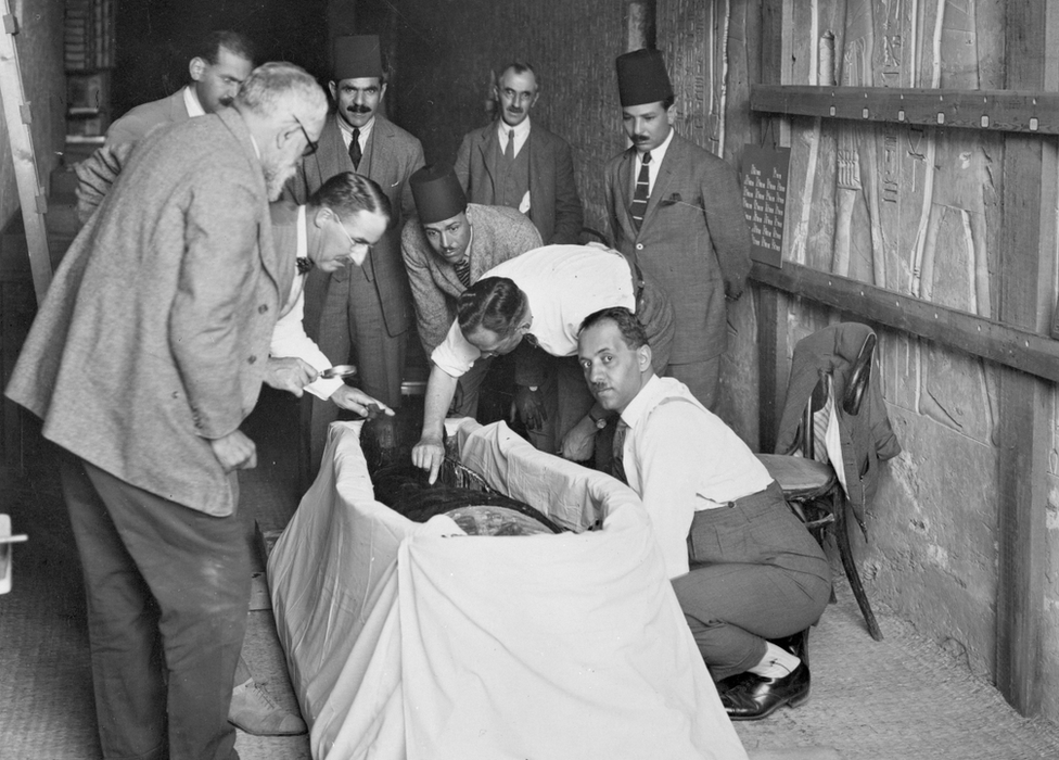 Diversos homens se curvam sobre o corpo de uma múmia; um deles tem um bisturi na mão e parece fazer uma incisão no corpo