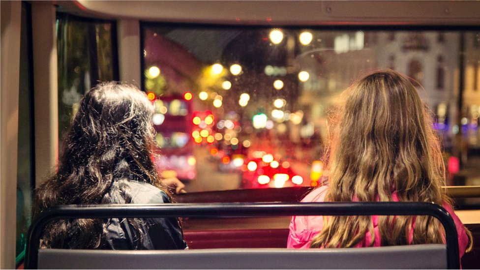 Fotografia colorida mostra duas mulheres em um ônibus sentadas de costas