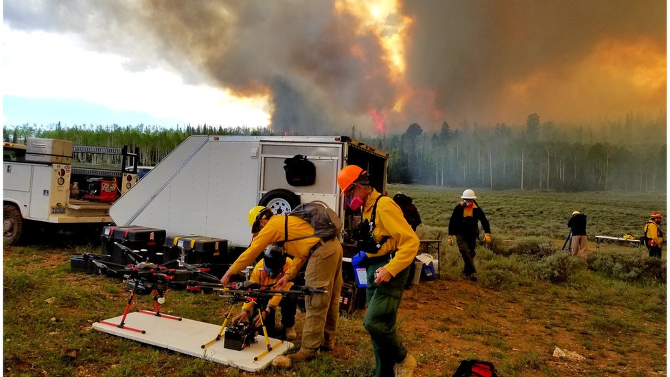 Cientistas paramentados e com equipamentos se preparam para fazer um drone decolar em frente a um incêndio florestal que solta muita fumaça