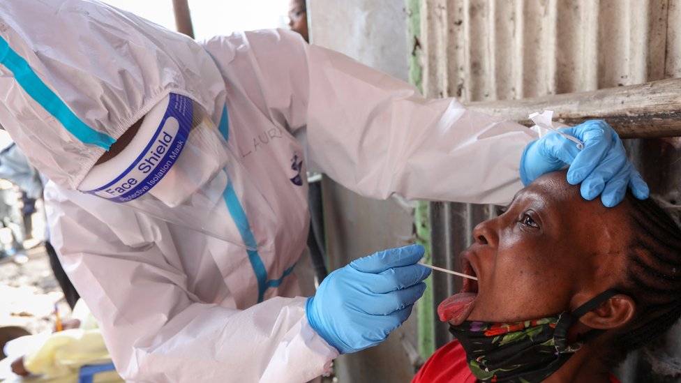 Кенийский медицинский работник в защитном снаряжении (слева) использует мазок из полости рта, чтобы взять образец у женщины во время массового тестирования на коронавирус SARS-CoV-2