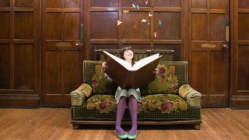 Una chica sentada en un sofá abre un libro gigante del que salen objetos de color