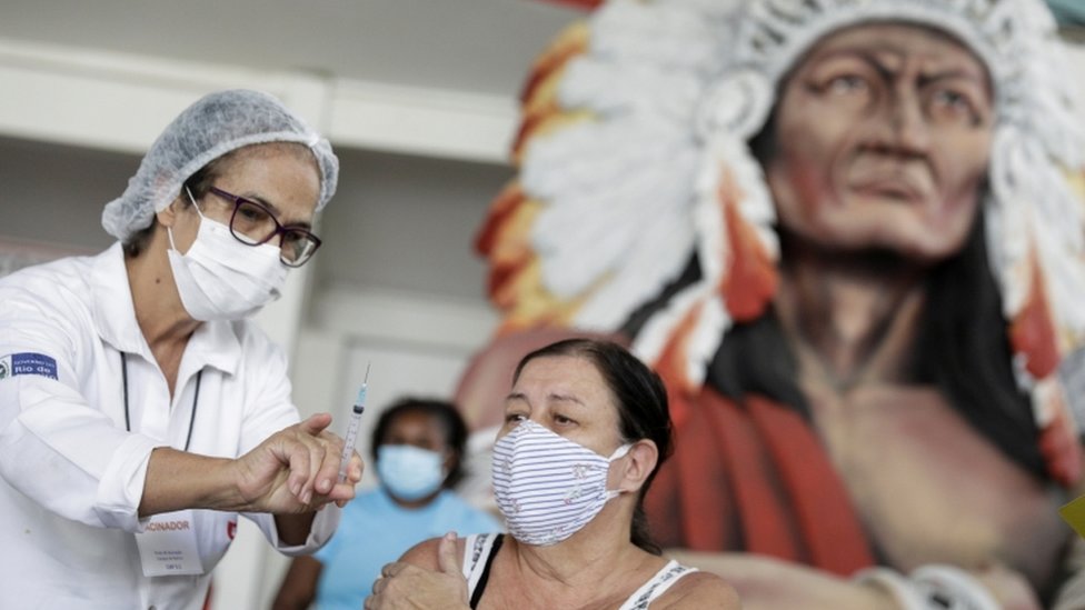 Profissional vacina mulher sentada; atrás, é possível ver grande escultura carnavalesca de um cacique