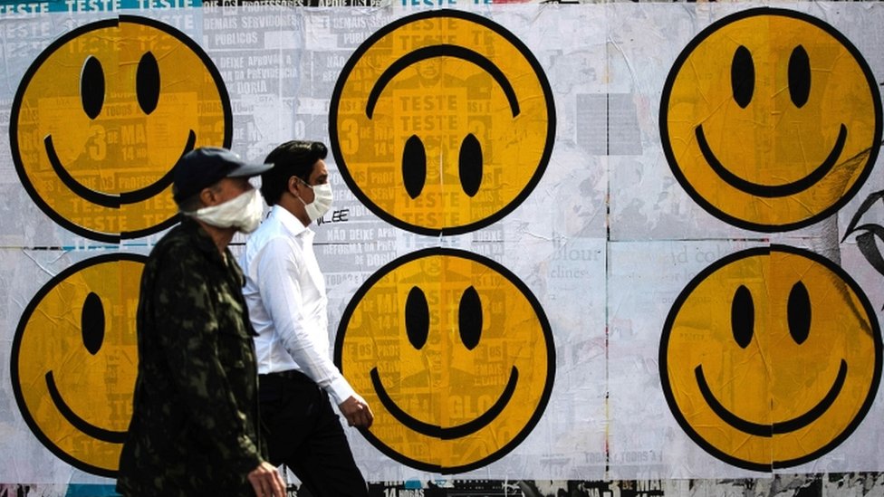 Dois pedestres com máscaras passam em frente a cartazes com carinhas felizes, uma delas de cabeça pra baixo