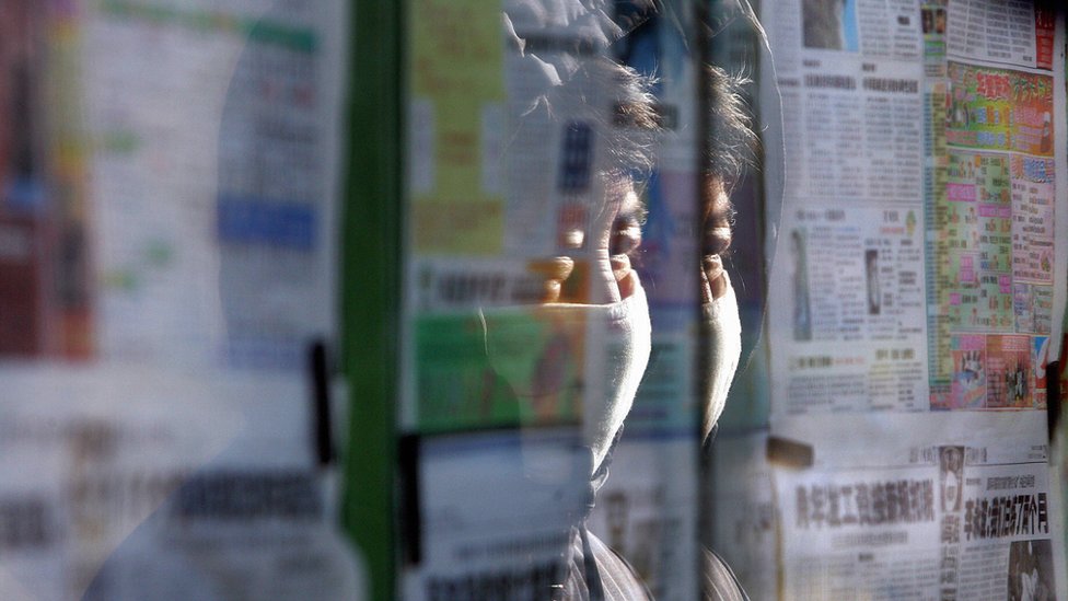 Женщина в маске читает новости из подконтрольных государству газет, вывешенных за стеклянными ограждениями на улице Пекина