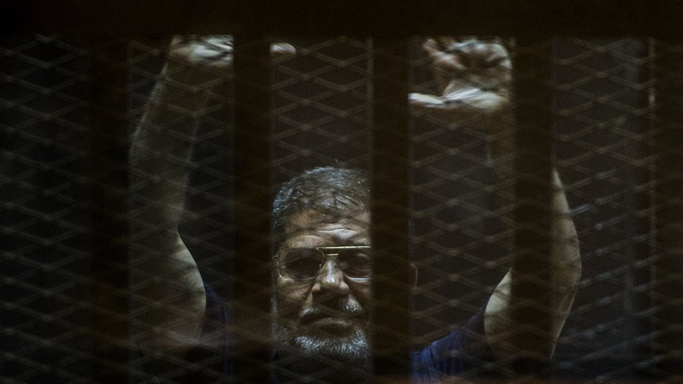Свергнутый президент Египта Мохамед Мурси жестикулирует из клетки обвиняемых, когда он присутствует на суде в полицейской академии на окраине столицы Каира