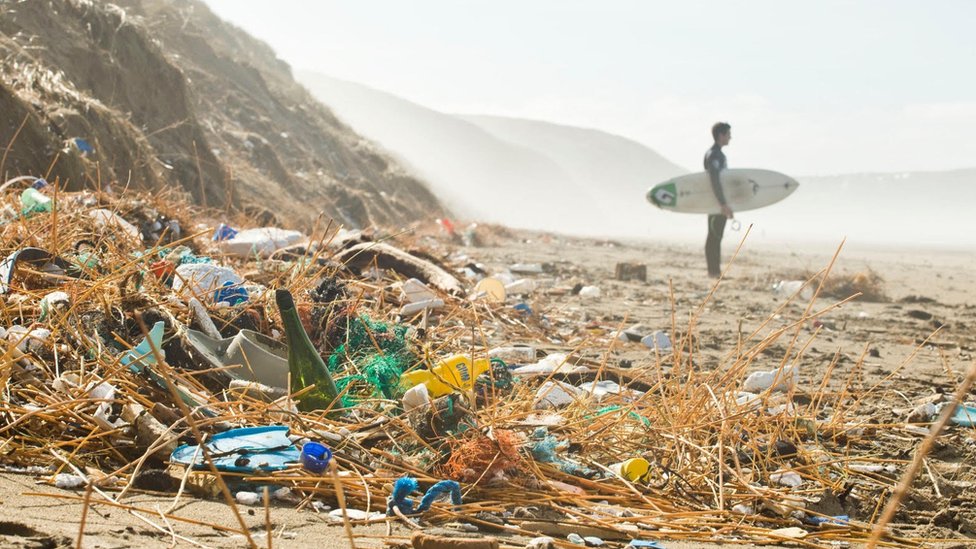 Серфер стоит возле холма пластикового мусора на пляже в Корнуолле