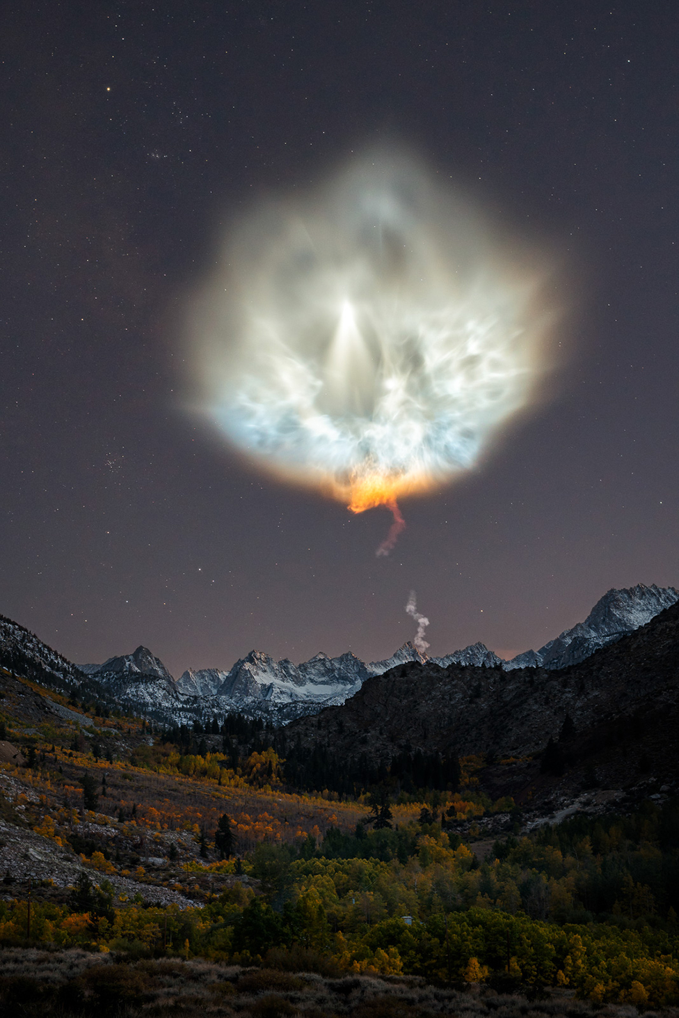 Пейзажная сцена, показывающая горы ночью с большим шлейфом выхлопных газов ракеты в небе