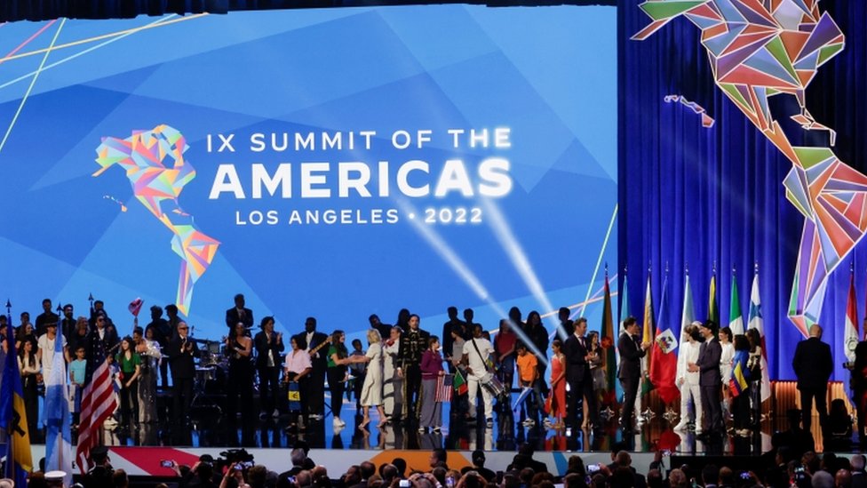 Dezenas de pessoas no palco, com telão mostrando símbolo da Cúpula das Américas