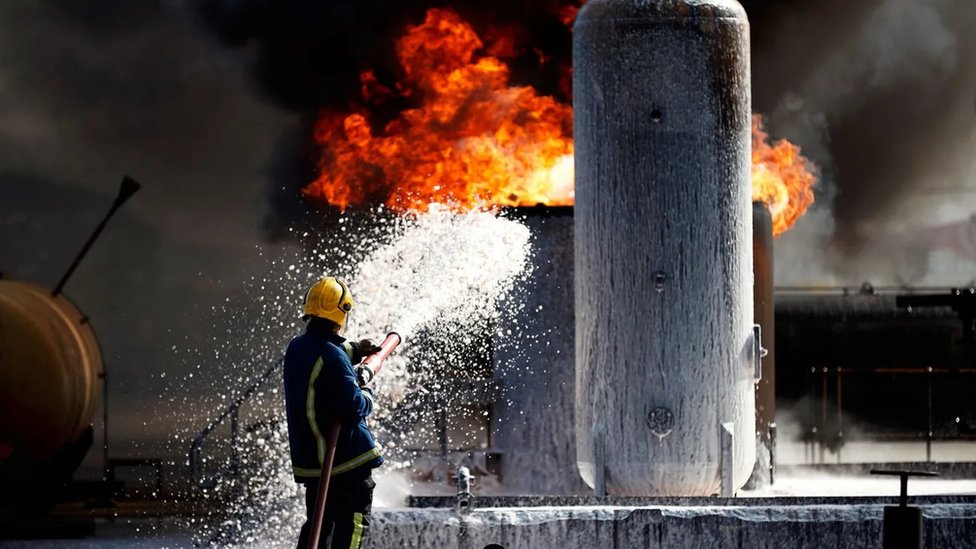 Fotografia colorida mostra bombeiro usando mangueira para apagar fogo em um imóvel que parece ser industrial