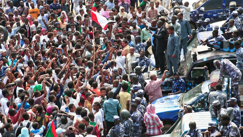 Суданский демонстрант машет руками, стоя на капоте автомобиля сил безопасности
