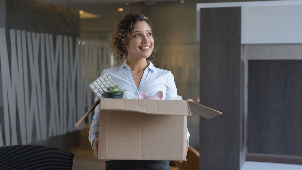 Una mujer sonríe mientras sostiene una caja llena de cosa de oficina.
