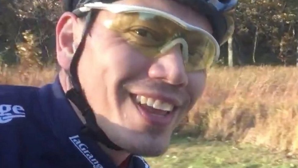 Tom Justice sorrindo e usando aparato de bicicleta com árvores ao fundo