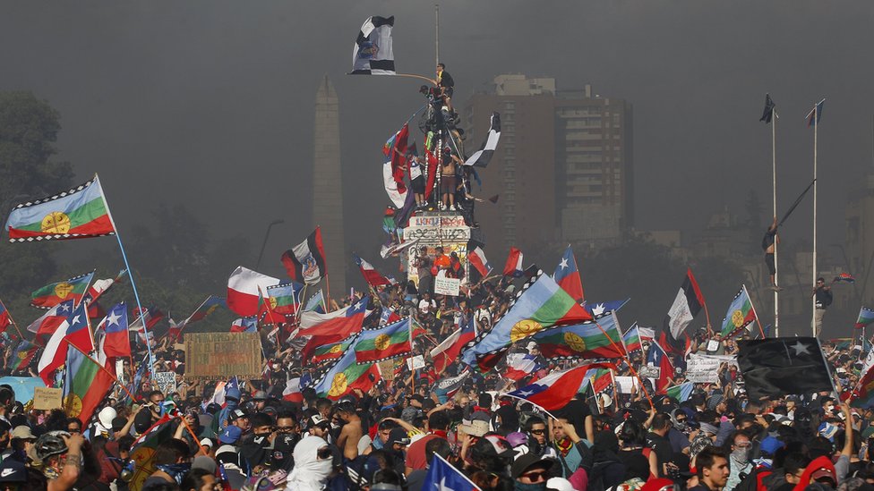 Протестующие выкрикивают лозунги и размахивают флагами Чили и народа мапуче во время национальной забастовки и всеобщей демонстрации, созванной различными профсоюзами трудящихся 12 ноября 2019 года в Сантьяго, Чили.