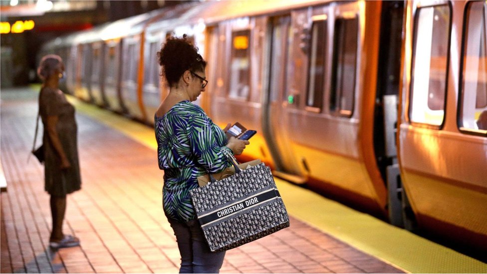 Fotografia colorida mostra mulher olhando o celular em uma estação de metro em frente a um vagão