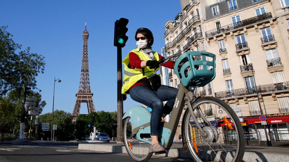 Женщина в маске едет на велосипеде по Парижу, за ее спиной запечатлена Эйфелева башня
