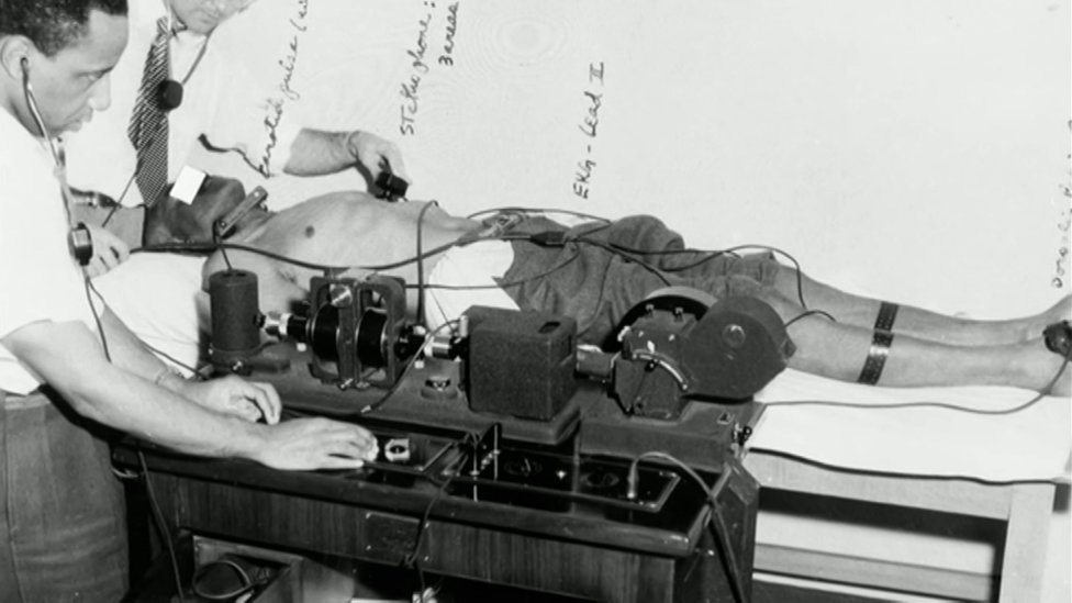 Foto em preto e branco mostra dois homens, um deles branco, usando aparelhos antigos para fazer experimentos em um homem negro deitado sem camisa