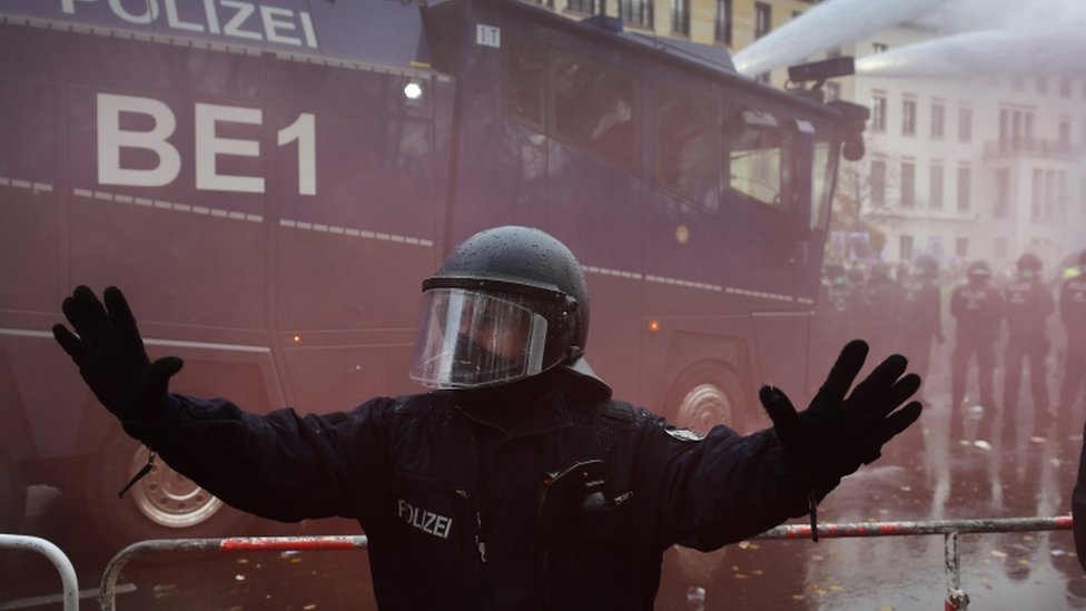 Полицейский поднимает руки, пока ОМОН с водометом разгоняет демонстрацию против ограничений, введенных в отношении немецкого коронавируса