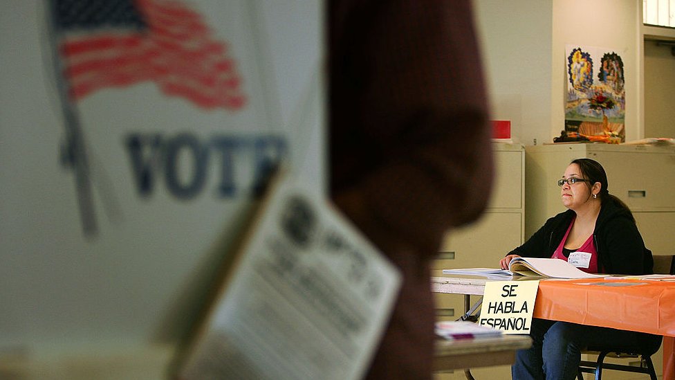 Una miembro de mesa de votación en Estados Unidos sentada en una mesa con un cartel que cuelga y dice Se habla español