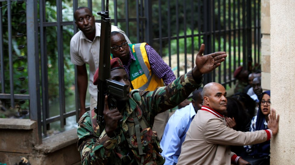 A member of the security forces gestures as people take cover at the scene where explosions and gunshots were heard at the Dusit hotel compound, in Nairobi, Kenya