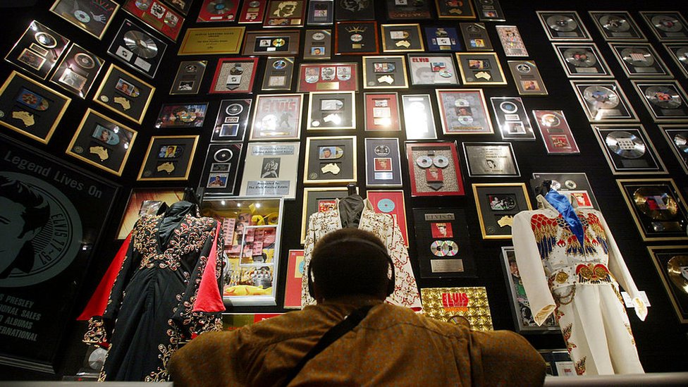 Uma pessoa olha para uma parede cheia de recordações de Elvis, como discos, discos de ouro e ternos
