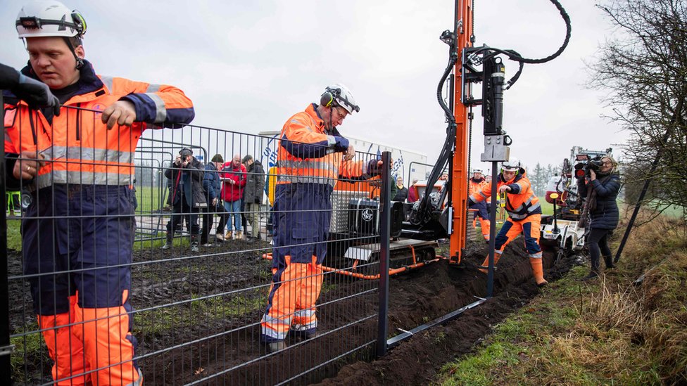 Radnici u narandžastim uniformama sastavljaju prve delove metalne ograde na granici Danse, dok ih snimatelji i fotografi slikaju