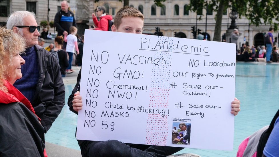 На субботней акции протеста проводится табличка с надписью «Никаких вакцин, никаких ГМО, никакой торговли детьми, никакой NWO»