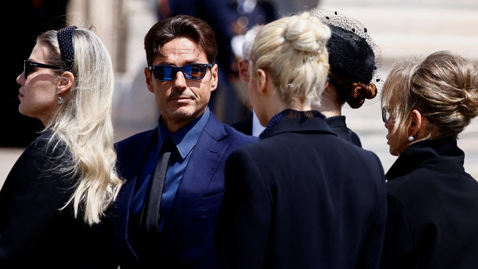 A group of five people visible at the funeral of Silvio Berlusconi. Pier Silvio is the only male visible. He is wearing dark glasses, a dark suit, and is looking towards the camera.