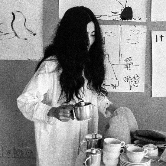 Fotografia em preto e branco mostra uma mulher de roupa branca e cabelos pretos compridos desarrumados servindo uma xícara de chá em frente a uma parede com desenhors feitos em papel