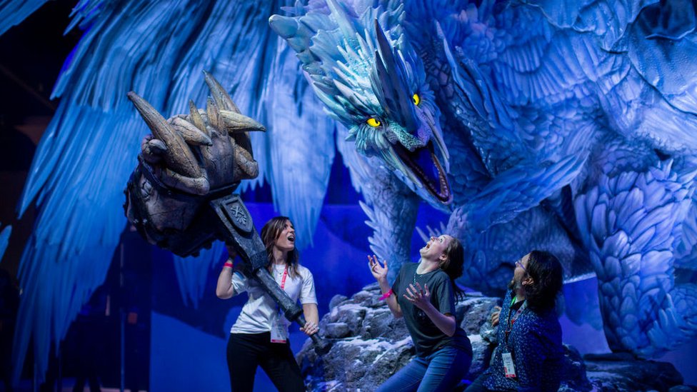 Три человека прячутся под гигантской скульптурой дракона, причем один из людей держит в руках комично негабаритный фантастический молот