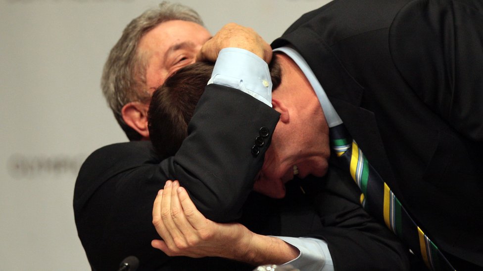 Sentado, Lula abraça Eduardo Paes, que se abaixa em direção do ex-presidente - ambos rindo em ambiente fechado