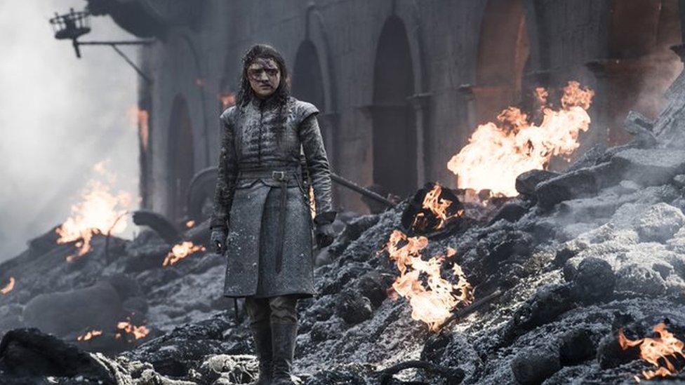 A scene from season 8 of Game of Thrones shows the character Arya Stark walking through burning rubble