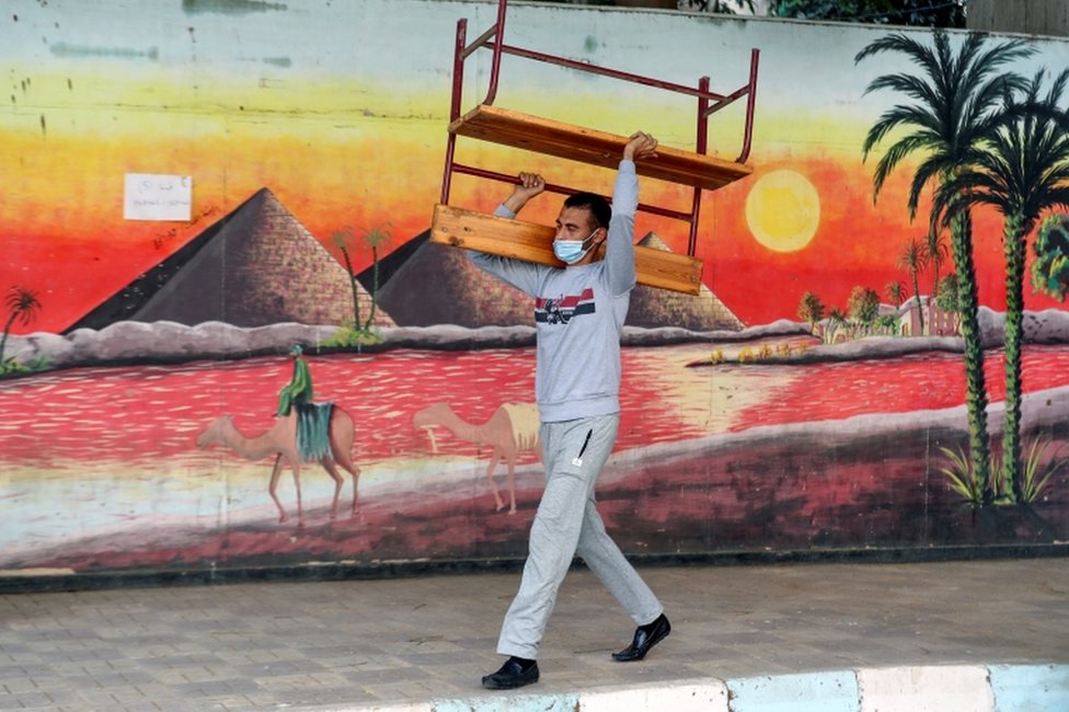 A man carries a bench outside a school, which has a mural painted on a wall.